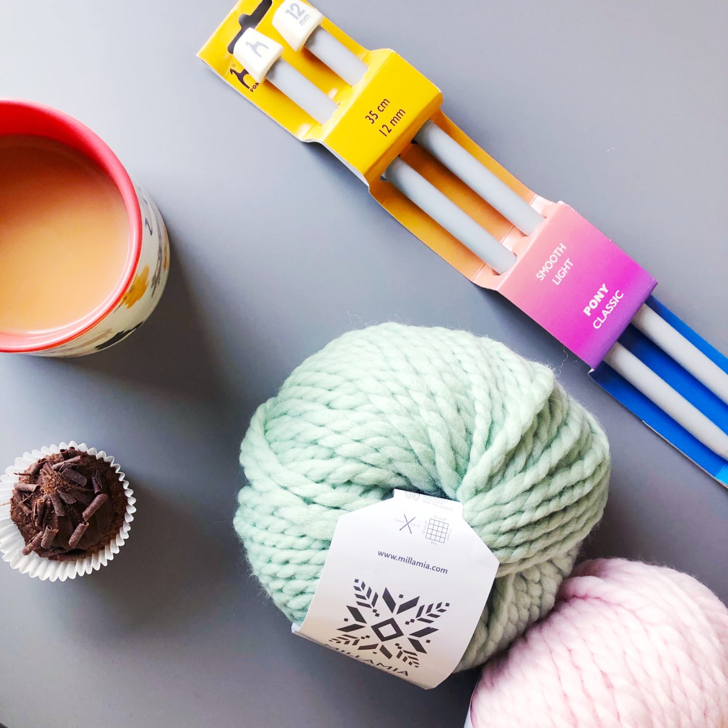 flatly of mint green and pale pink Millamia balls of wool with size 12 knitting needles, a cup of tea and a mini cupcake on a grey background